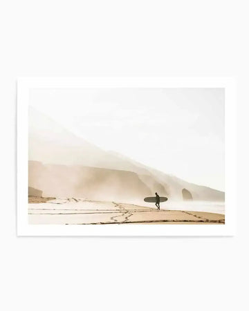 Framed photograph of a lone surfer walking along a misty beach at sunrise, with a natural wood frame and white border. The composition features a silhouette of a person carrying a surfboard, walking on wet sand with footprints, against a hazy background of rolling hills and a single rock formation. The scene is bathed in soft, warm light, creating a serene and atmospheric mood.