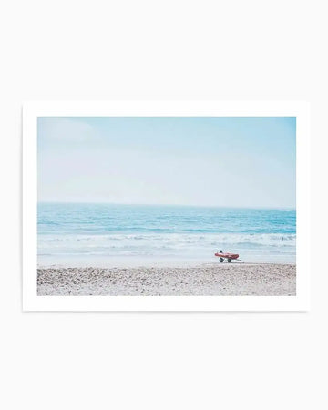 Framed art print featuring a serene coastal photograph with a light blue matted border. The image shows a sandy beach in the foreground, calm turquoise ocean waters in the middle ground, and a pale blue sky above. A person carrying a red surfboard walks along the wet sand near the water's edge, with gentle waves and a few distant swimmers visible. The photograph has a soft, minimalist aesthetic with cool coastal tones of pale blue, turquoise, cream, and sandy beige. The print is mounted in a white frame.