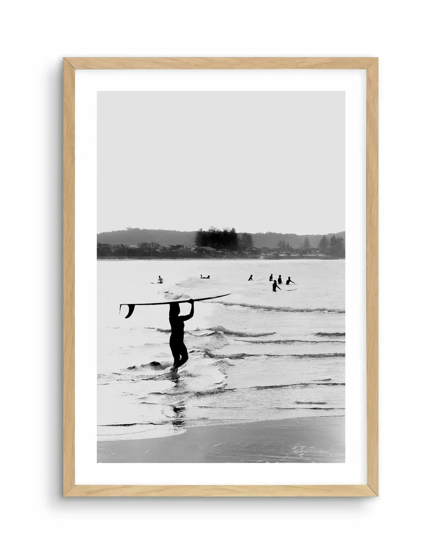 Black and white framed art print showing a surfer in a wetsuit carrying a surfboard overhead while walking through shallow ocean waves. In the background, other surfers are visible in the water with rolling waves, a tree-lined shoreline, and hills under an overcast sky. The photograph has a documentary-style aesthetic with high contrast and grainy texture typical of film photography. Natural wood-toned frame with white border surrounding the vertical composition.