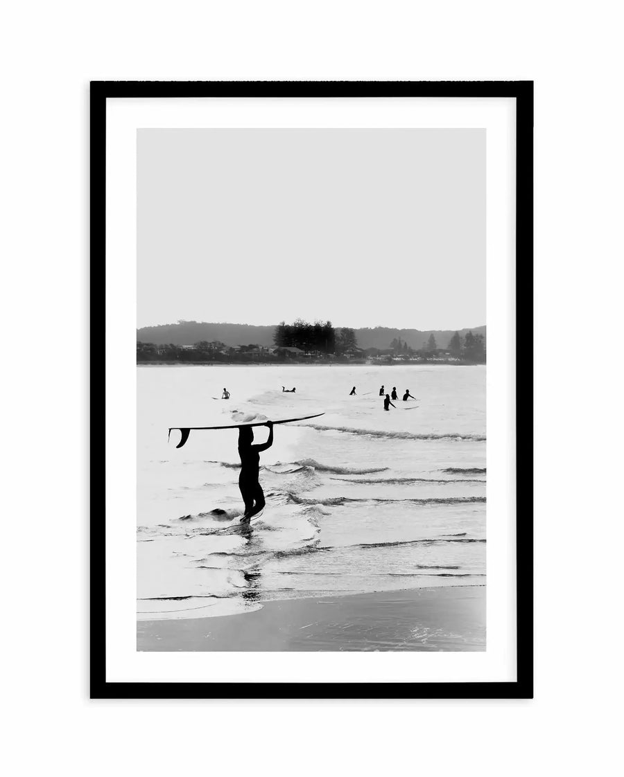 Black and white photograph framed in a black frame with white border, depicting a surfer in a wetsuit carrying a surfboard overhead while walking through shallow ocean waves. In the background, other surfers are visible in the water, with rolling hills and trees lining the distant coastline under an overcast sky. The composition captures a moody, atmospheric beach scene with strong contrast between the dark silhouette of the surfer and the lighter tones of the water and sky.
