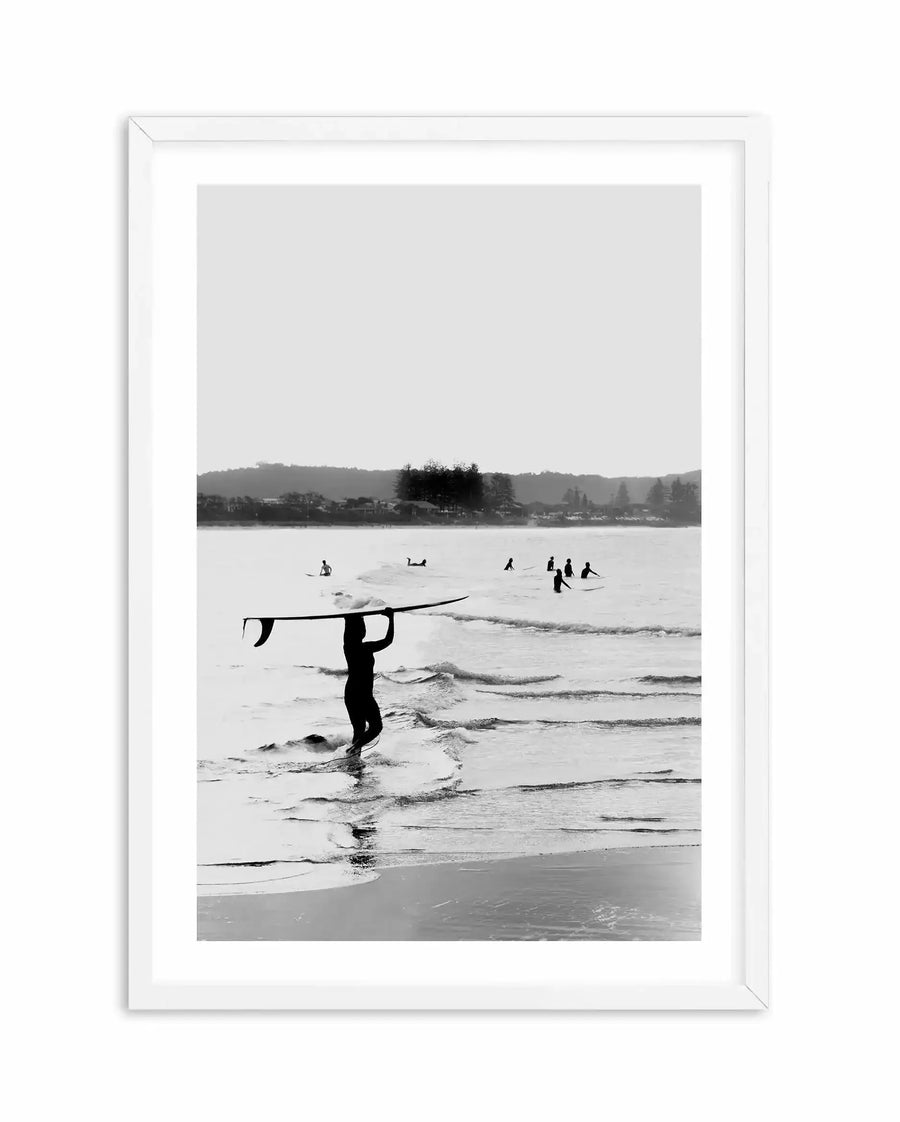 Black and white photograph printed on paper, displayed in a white wooden frame with white border. The image shows a surfer in a wetsuit carrying a surfboard overhead, walking through shallow ocean waves. Multiple other surfers are visible in the water in the background. Rolling hills and coastal buildings line the distant shoreline beneath an overcast sky. The composition captures a moody, contemplative beach scene with strong contrast between the dark silhouetted figure and the lighter water and sky.