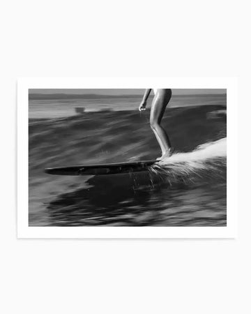 Black and white photograph of a surfer girl in motion on a wave in Malibu. The dynamic composition captures her legs and surfboard from a low angle as she rides an active wave, with water spray and motion blur conveying speed and energy. The monochromatic film photography style emphasizes contrast between her pale skin and the dark water and board. The image is printed on white paper with a clean white border, presented as a framed art print.