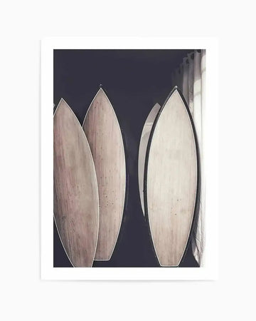 Framed photograph of four surfboards leaning against a dark wall, with a window and curtains visible on the right. The surfboards are light wood with dark edges, and the image has a muted, slightly desaturated colour palette.