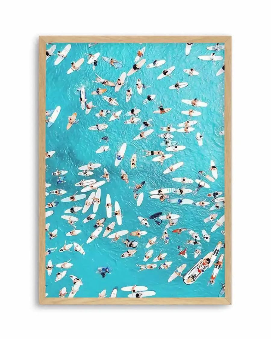 Framed aerial photography print of a vibrant turquoise ocean filled with numerous surfers on white and light-colored surfboards, viewed from directly above. The composition captures the surfers in various positions, some paddling, some resting, and some appearing to ride small waves, creating a lively and dynamic scene. The print is displayed in a natural oak frame with a white border.