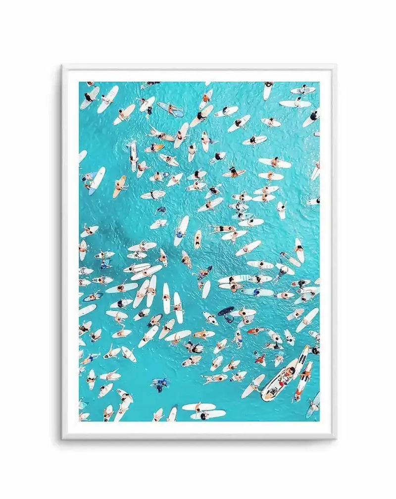 Framed aerial photograph of a vibrant turquoise ocean filled with numerous surfers and their white surfboards, viewed from directly above. The composition captures the dynamic movement of people and boards scattered across the water, creating a lively and colourful scene. The print is displayed in a white frame with a white border.