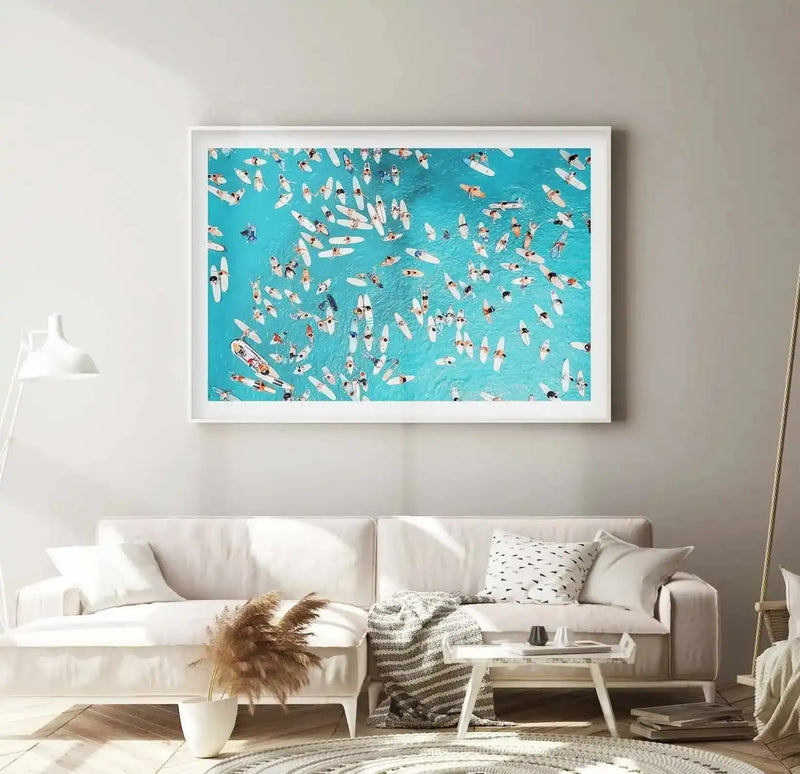 Lifestyle shot of a minimalist living room with a large framed art print displayed above a white sofa. The print is a vibrant aerial photograph of numerous surfers and paddleboarders on a bright turquoise ocean, with white surfboards scattered across the water. The room features a white floor lamp, a textured cream throw blanket, dried pampas grass in a white vase, and a small white coffee table with books.