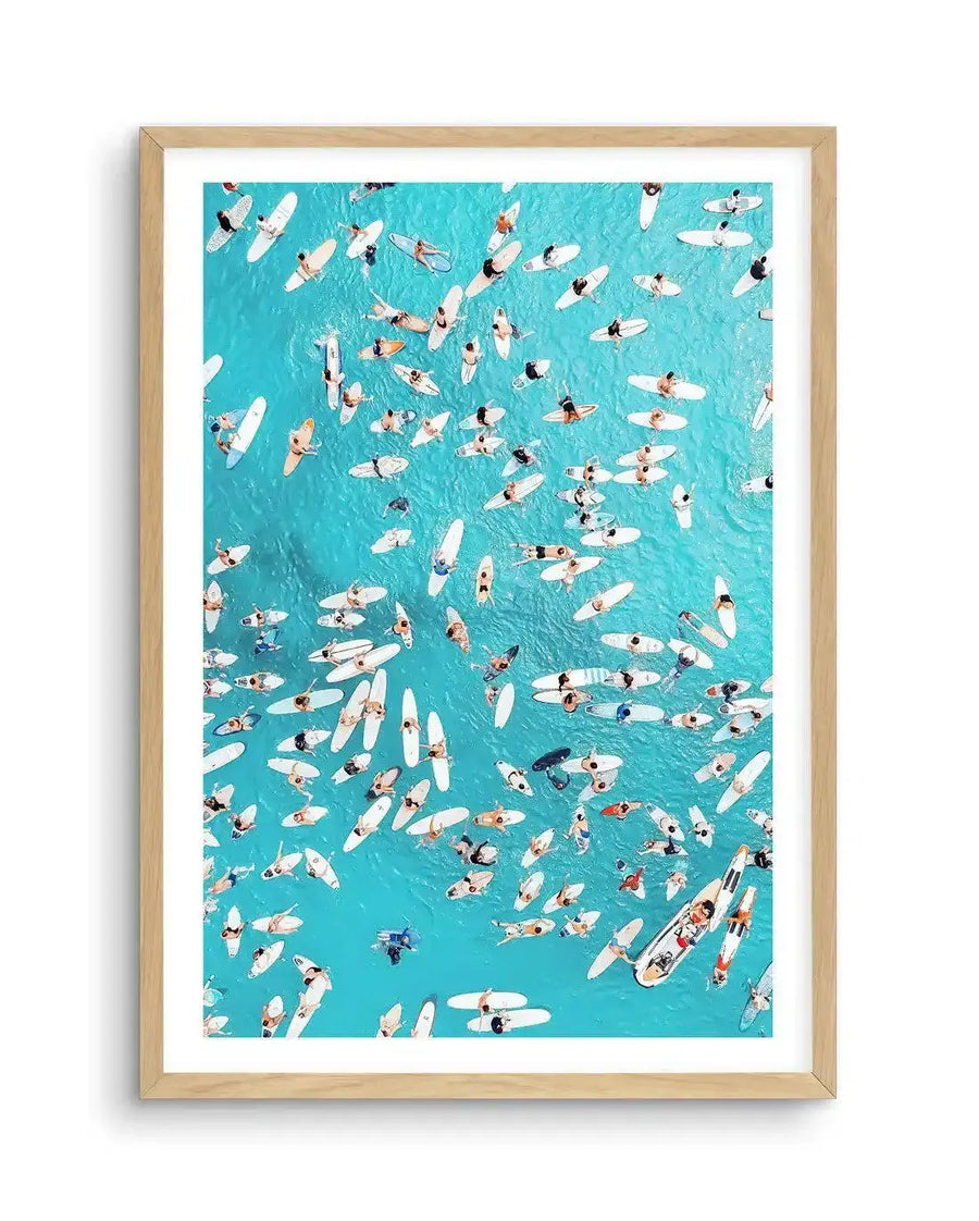 Framed aerial photograph of a large group of surfers and their surfboards floating on a clear turquoise ocean, with a natural wood frame and white border. The composition captures the vibrant blue water and the scattered white surfboards and swimmers from a bird's-eye view.
