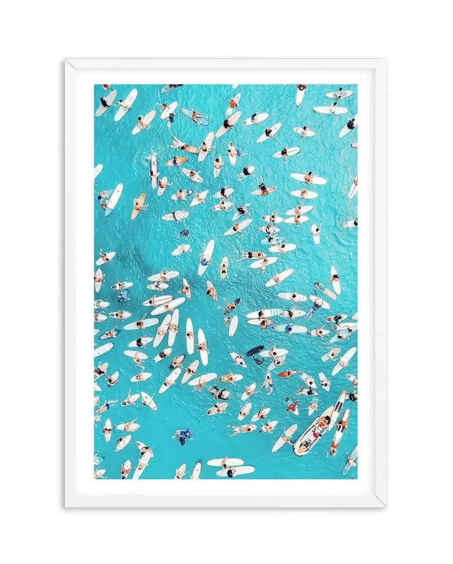 Framed aerial photograph of a vibrant turquoise ocean filled with numerous surfers and their white surfboards, displayed in a white frame with a white border. The composition captures the surfers from directly above, creating a pattern of white boards and human figures against the bright blue water.