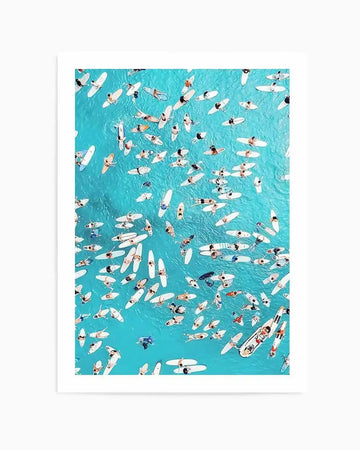 Aerial photography art print of a vibrant turquoise ocean filled with numerous surfers on white surfboards, creating a dynamic and crowded scene. The composition captures the surfers from above, some paddling, some resting, and some in the water next to their boards, against the bright blue water.