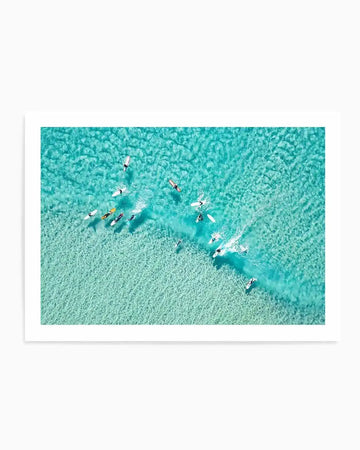 Aerial photograph art print of surfers in clear turquoise water, with a white border. The composition shows multiple surfers on white and light-coloured boards, some paddling, some standing, creating small wakes in the vibrant blue-green ocean. The water reveals a sandy bottom with ripple patterns.