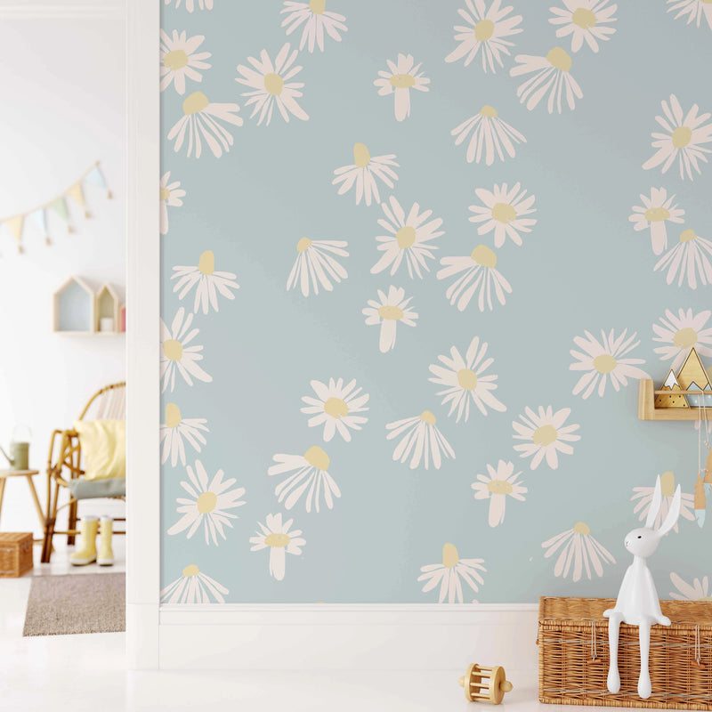 Lifestyle shot of a child's room featuring Sweet Daisy Flower Wallpaper. The wallpaper has a repeating pattern of hand-painted white daisy flowers with pale yellow centers on a soft blue-green background. The room shows a white floor and baseboards, with a glimpse into another room featuring a rattan chair, yellow cushion, and decorative bunting. A white rabbit figurine sits on a wicker basket in the foreground.