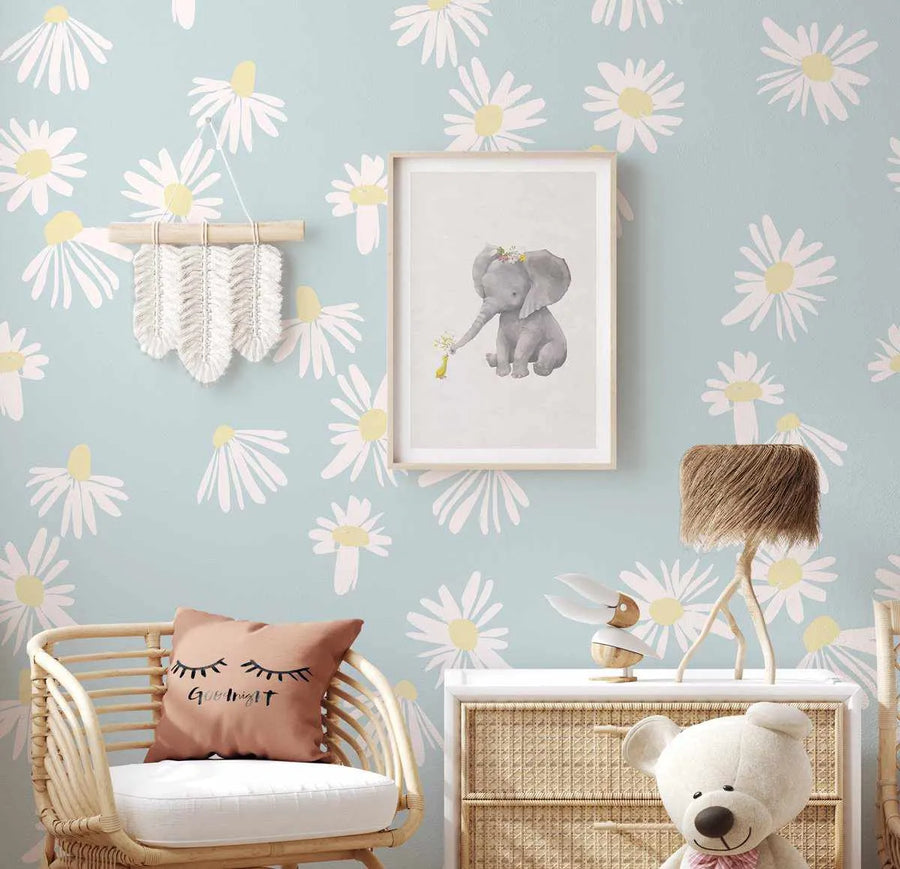 Lifestyle shot of a child's bedroom featuring Sweet Daisy Flower Wallpaper. The wallpaper has a repeating pattern of hand-painted white daisy flowers with pale yellow centers on a soft blue background. The room includes a framed art print of a baby elephant with a flower crown holding a small yellow bird, a rattan chair with a "Goodnight" pillow, a macrame wall hanging, a wooden dresser with a woven texture, a wooden bird figurine, a lamp with a textured brown shade, and a white teddy bear.