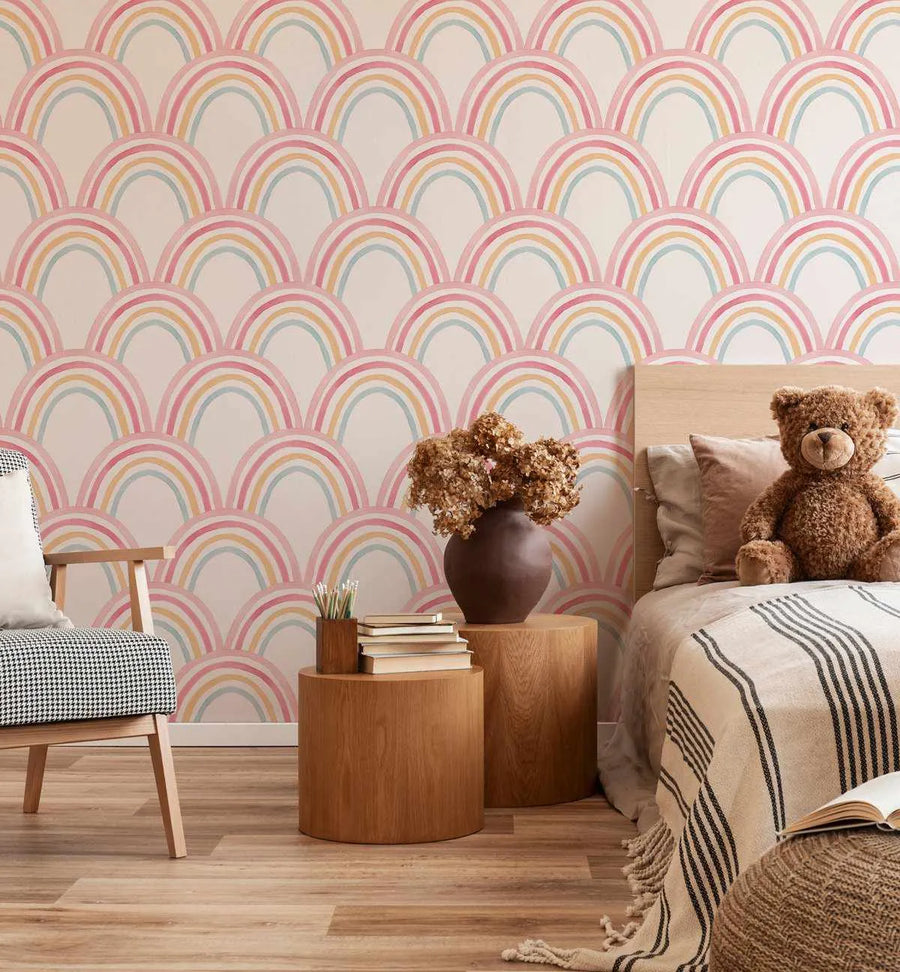 Sweet Rainbows wallpaper featuring a repeating pattern of hand-painted arched rainbows in soft pink, pale yellow, and light blue on a cream background, installed in a child's bedroom. The room includes a light wood bed with a teddy bear, a wooden armchair with a black and white cushion, and two round wooden side tables with books and a vase of dried flowers.