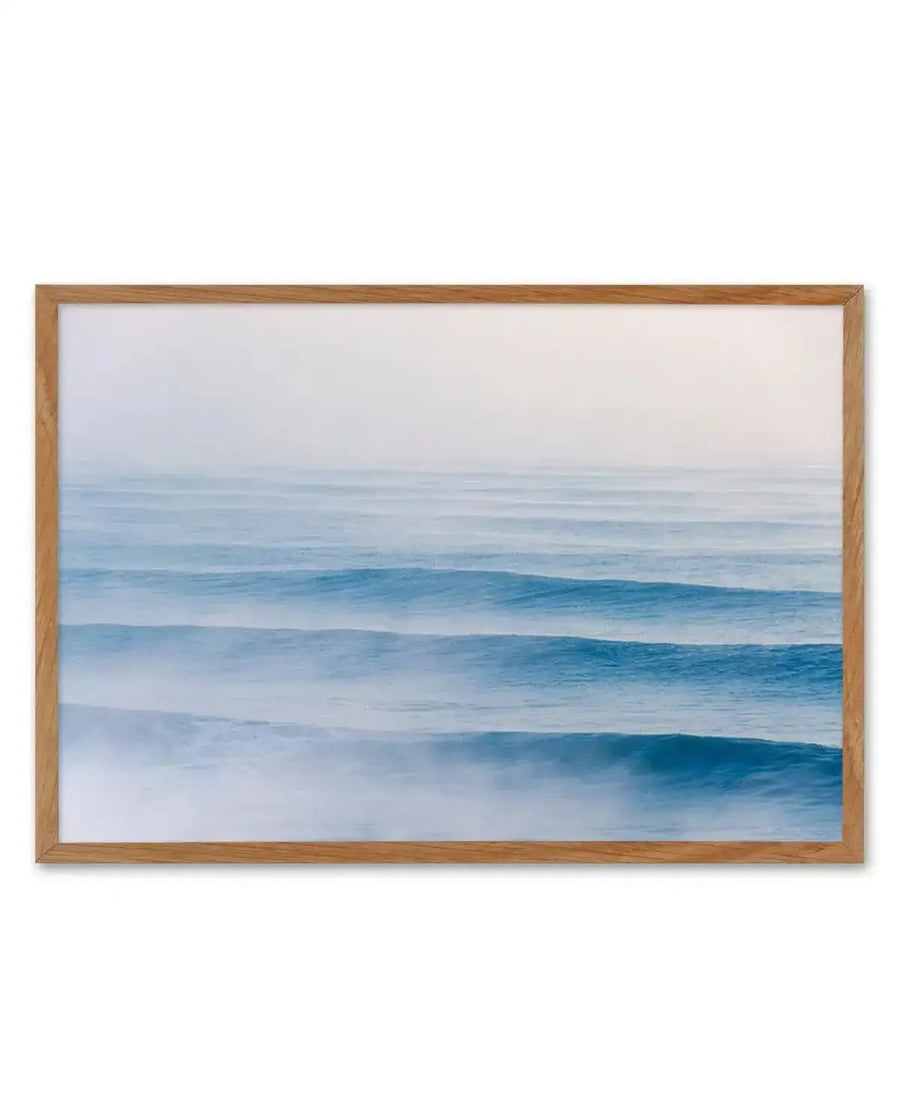 Framed photography art print featuring a serene ocean scene with multiple parallel lines of gentle waves rolling towards the viewer under a soft, hazy sky. The water transitions from a deep blue in the foreground to a lighter, misty blue towards the horizon, with a light blue-grey sky blending seamlessly into the distant water. The print is displayed in a natural oak frame with a white border.