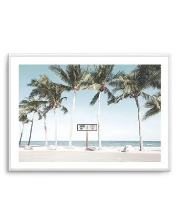 Framed photography art print featuring a tropical beach scene with a light blue sky and calm ocean, with a white frame and white border. The composition shows a row of palm trees on a white sand beach, with a wooden sign in the foreground pointing left for "SWIM" and right for "SURF".