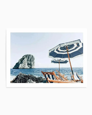 Framed photograph of a serene beach scene with a large rock formation in the distance, natural wood frame and white border. The foreground features two blue and white striped beach umbrellas and several matching deck chairs on a rocky shore, with calm blue ocean water stretching to the horizon under a clear, pale blue sky.