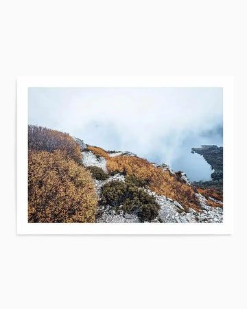 Framed photograph of a misty mountain landscape with a lake, featuring autumn foliage in shades of golden yellow and deep orange against a backdrop of grey and white clouds. The composition shows rocky terrain and dark green shrubs in the foreground, with a serene lake partially visible through the mist on the right side.