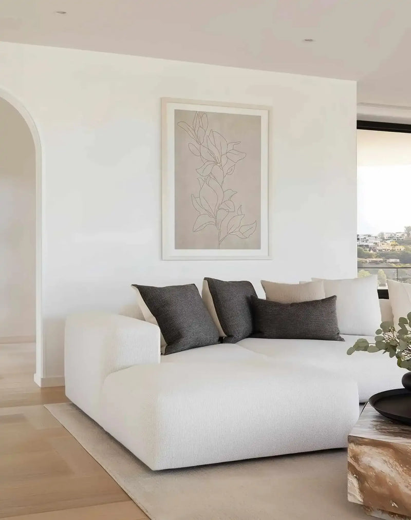 Large framed art print displayed above a white modular sofa in a minimalist living room. The print features a single-line illustration of a botanical branch with several leaves in a light grey against a warm beige background, with a white frame and white border. The room includes dark grey and beige throw pillows, a light-coloured rug, and a view of a city on a hill through a large window.