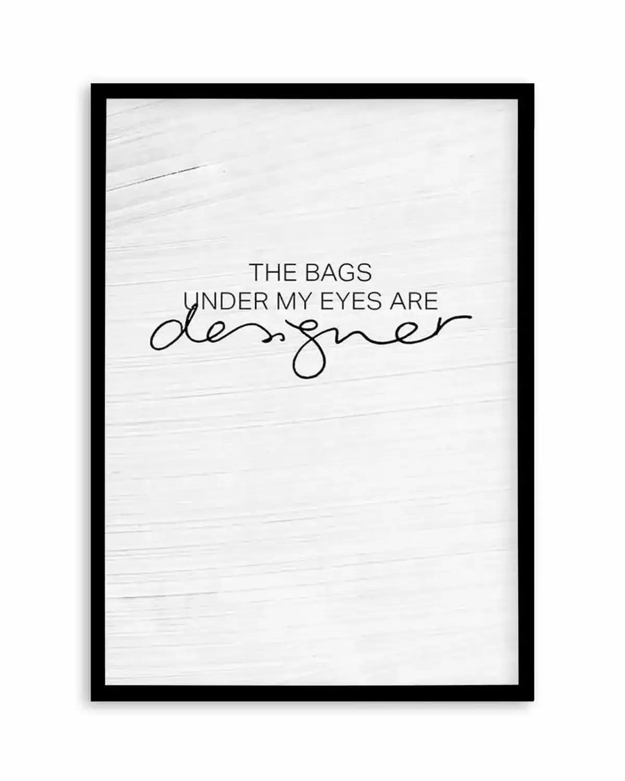 Framed art print with black wood frame featuring a white textured background with horizontal wood grain pattern. Text reads "THE BAGS UNDER MY EYES ARE" in sans-serif font, with "designer" written in flowing, cursive script below. Minimalist black and white design with humorous sentiment about tired appearance.