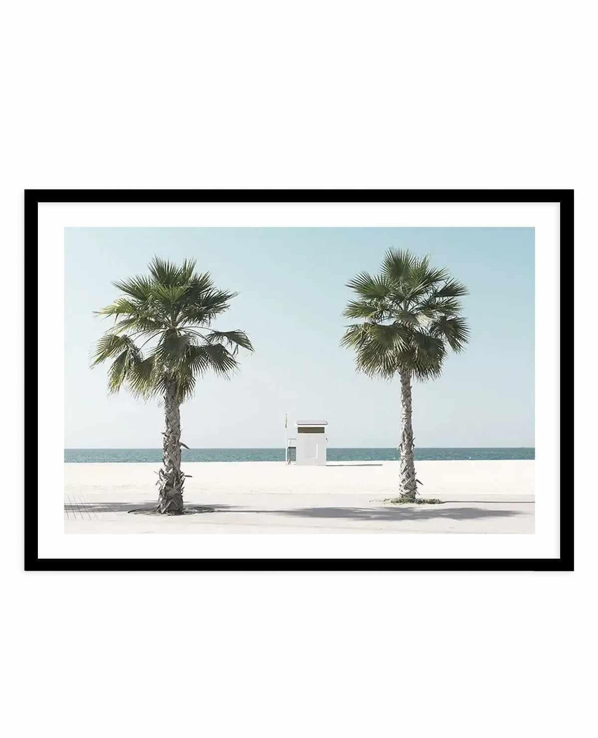 Framed art print featuring a minimalist beach photograph. Two palm trees with green fronds stand on pale sand, flanking a small white lifeguard station in the center. Clear turquoise water and pale blue sky form the background. The photograph has a serene, symmetrical composition with soft natural lighting and muted tones. Black frame with white border surrounding the image.