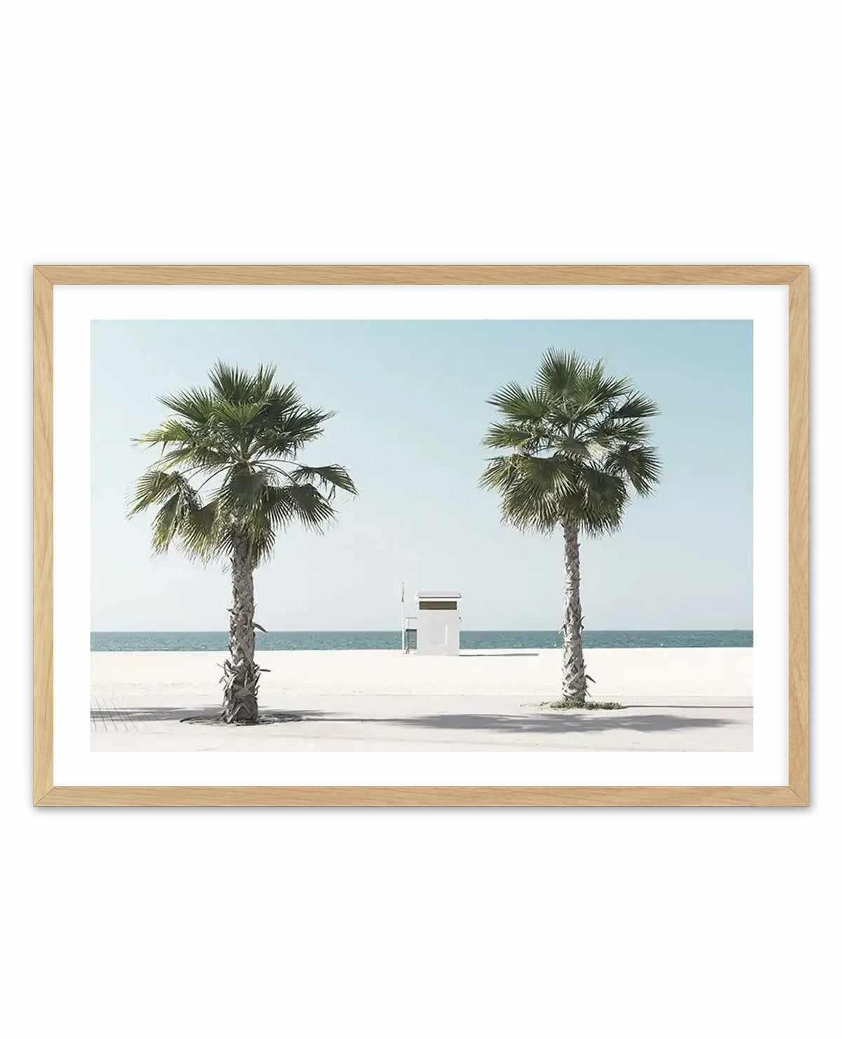 Framed art print featuring a minimalist beach scene photograph with a natural wood frame and white border. The image shows two tall palm trees with green fronds flanking a small white lifeguard station on a sandy beach. Clear turquoise ocean and pale blue sky create a serene, symmetrical composition. The photograph has a clean, contemporary aesthetic with soft lighting and muted tones of cream, sage green, and pale aqua.