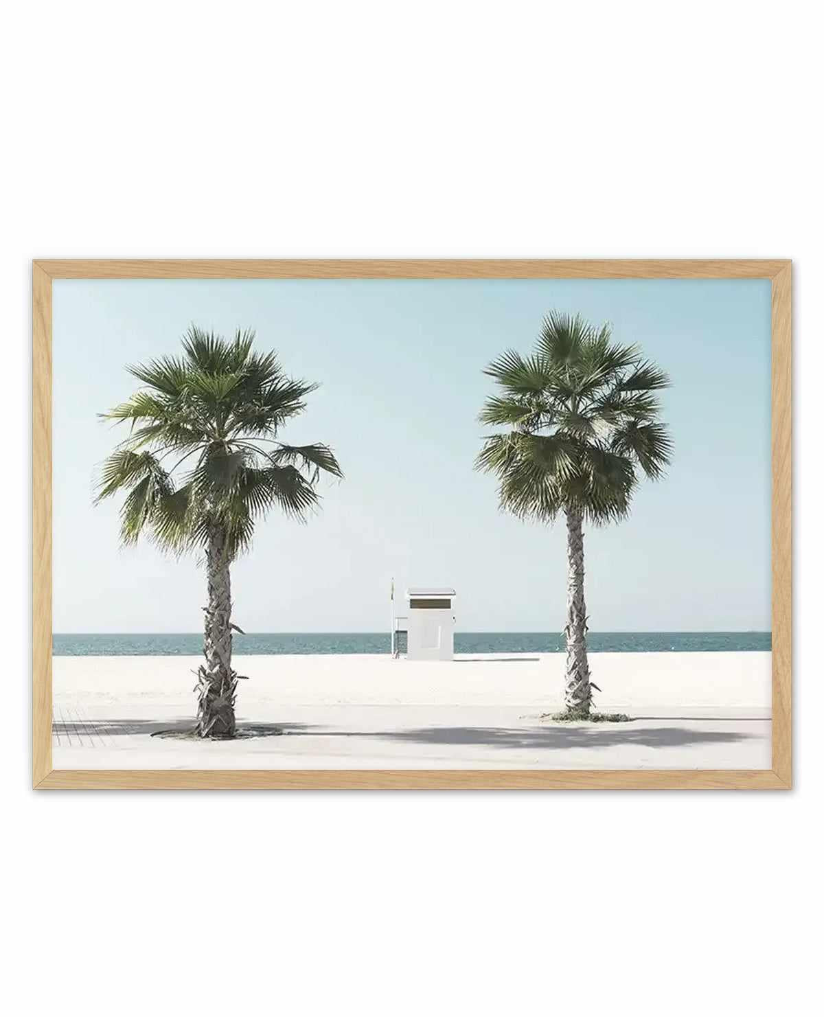 Framed art print featuring a minimalist beach scene with two tall palm trees flanking a small white lifeguard station on pale sand. Clear turquoise water and soft blue sky create a serene, horizontal composition. Natural wood frame with white border. Photographic style with muted, coastal colour palette of cream, sage green, and seafoam blue tones.