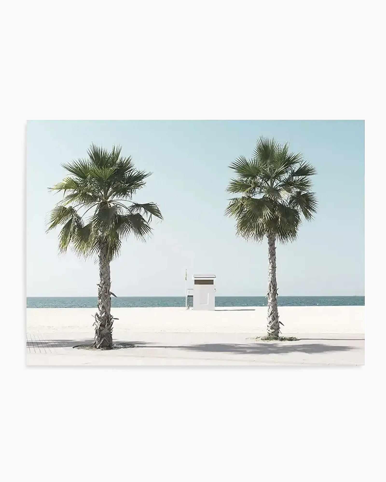 Minimalist beach scene art print featuring two palm trees with fan-shaped fronds flanking a small white lifeguard station centered on a wide sandy beach. Calm turquoise ocean and pale blue sky create a serene, symmetrical composition. The photograph-style image uses a soft, muted color palette of cream, sage green, and seafoam tones. Presented as a horizontal rectangular print with white border and light wood frame.