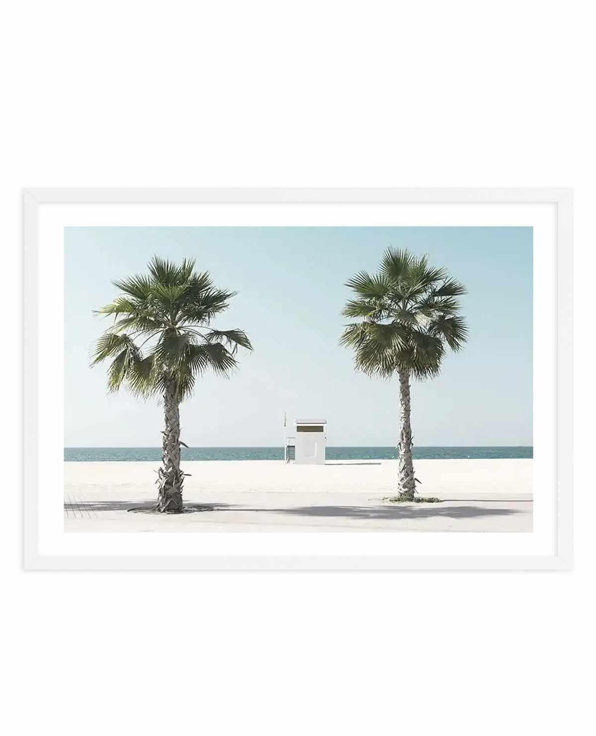 Framed art print featuring a serene beach scene with two tall palm trees flanking a small white lifeguard station. The composition shows a wide sandy beach in the foreground with calm turquoise ocean and pale blue sky in the background. The photograph has a minimalist aesthetic with clean lines and soft, muted tones of cream, sage green, and seafoam blue. The print is mounted in a white frame with white border.