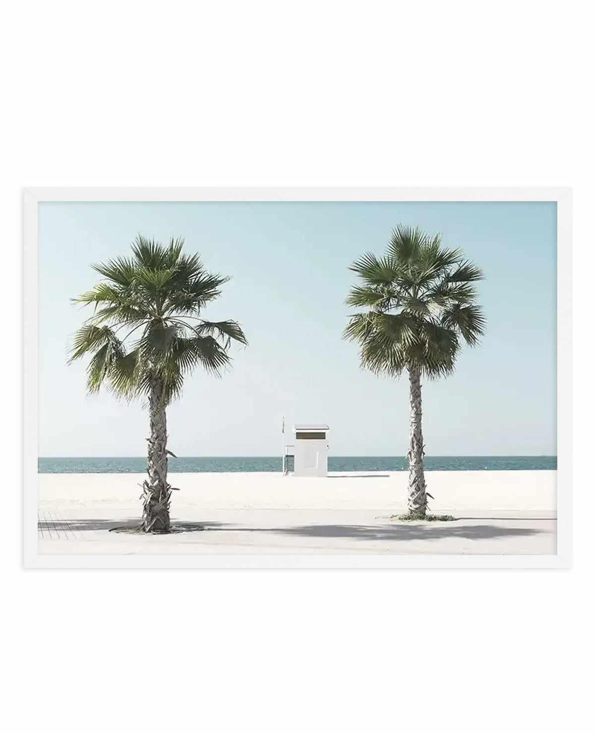Framed art print featuring a minimalist beach scene with two tall palm trees flanking a small white lifeguard station on pale sand. Clear turquoise water and soft blue sky create a calm, symmetrical composition. The photograph has a clean, contemporary aesthetic with soft natural lighting and subtle shadows. White frame with white border.