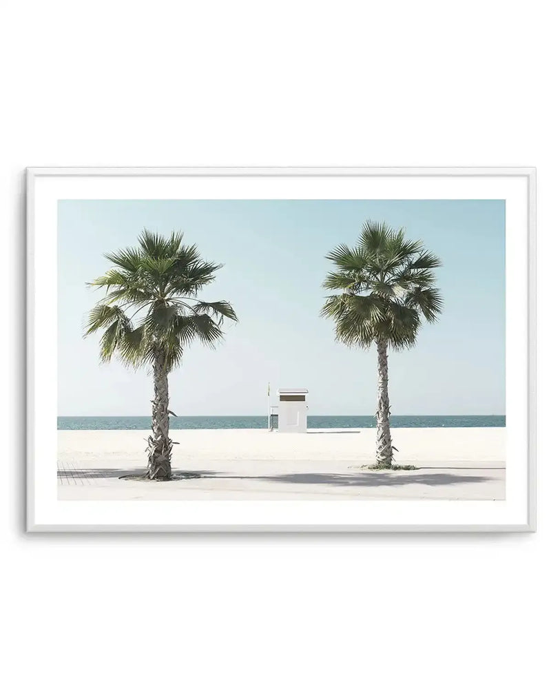 Framed photography art print featuring a tranquil beach scene with two palm trees and a lifeguard stand, displayed in a white frame with white border. The composition shows two tall palm trees with green fronds on a light sandy beach, with a small white lifeguard stand in the distance near the calm blue ocean under a clear pale blue sky.