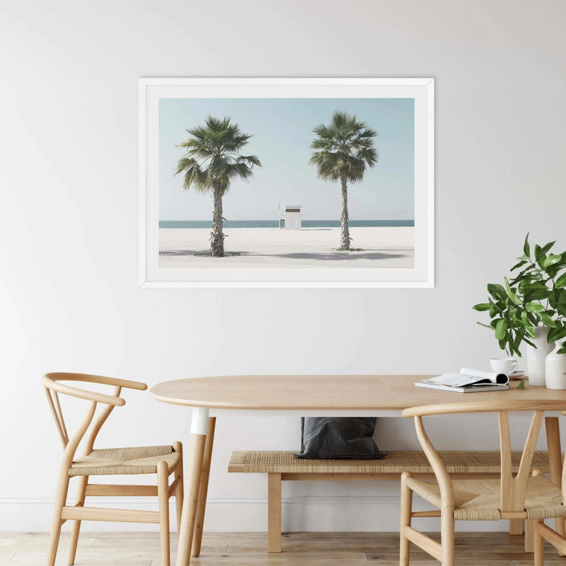 Lifestyle shot featuring a framed art print of a beach scene displayed above a wooden dining table in a bright, minimalist room. The print features two tall palm trees on a white sand beach with a small white lifeguard hut in the distance, and calm turquoise ocean under a pale blue sky. The art print has a white frame and white border. The room includes two light wood chairs with woven seats, a matching wooden bench with a dark grey cushion, and a vase of green foliage on the table.