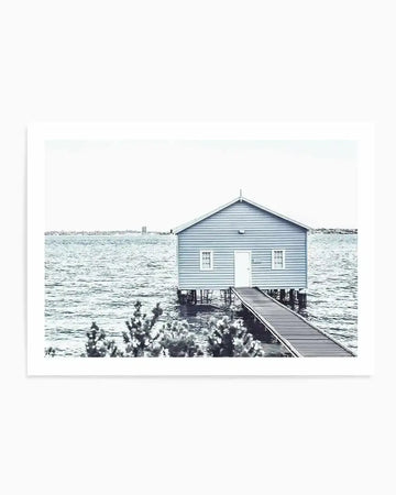 Framed black and white photograph of a boathouse on stilts over calm water, with a wooden pier leading to its white door. The boathouse has light blue siding and two white-framed windows on either side of the door. The water reflects a pale sky, and a distant city skyline is visible on the horizon. The foreground features blurred foliage.