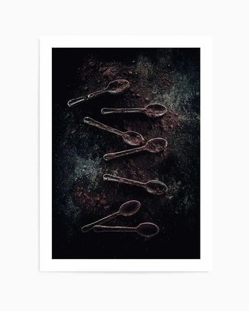 Framed dark and moody photograph of six chocolate-covered spoons arranged diagonally on a textured, dark background with scattered cocoa powder. The spoons appear to be made of clear glass or plastic, with the chocolate coating creating a rich, dark brown and reddish-brown hue. The background has a mottled, almost cosmic appearance with subtle specks of lighter colour, and the overall composition is dramatic and atmospheric.