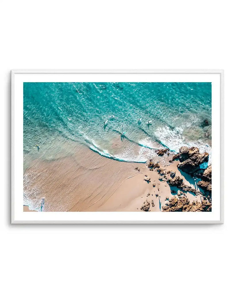 Framed aerial photograph of a coastal beach scene with a white frame and white border. The composition features surfers in turquoise water with gentle waves breaking on a sandy beach, and scattered rocks along the shore.