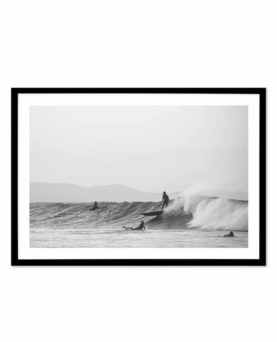 Framed black and white photograph of a surfing scene with a black frame and white border. The composition features five surfers in the ocean, with one actively riding a breaking wave, and mountains visible in the hazy background.
