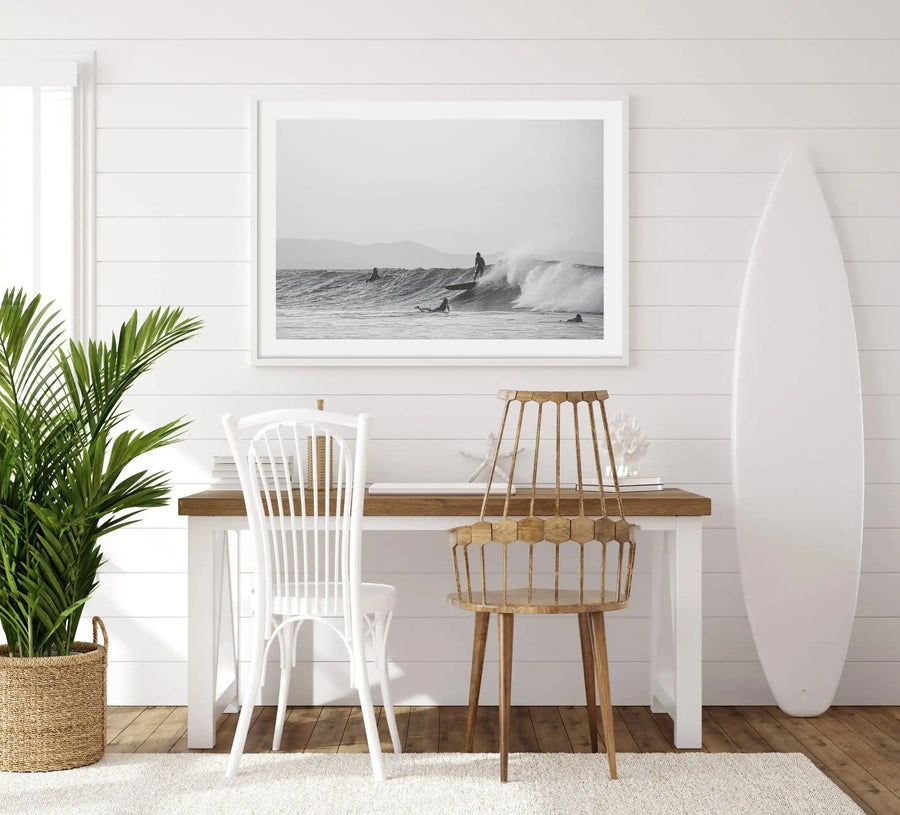 Lifestyle shot of a home office with a framed black and white photograph of surfers in the ocean, displayed above a wooden desk. The print features a large wave crashing with a surfer riding it, and other surfers paddling nearby, with mountains in the background. The room has white shiplap walls, a light wood floor with a cream rug, a large green plant in a woven basket, a white wooden chair, and a light wood chair with a woven back. A white surfboard leans against the wall on the right.