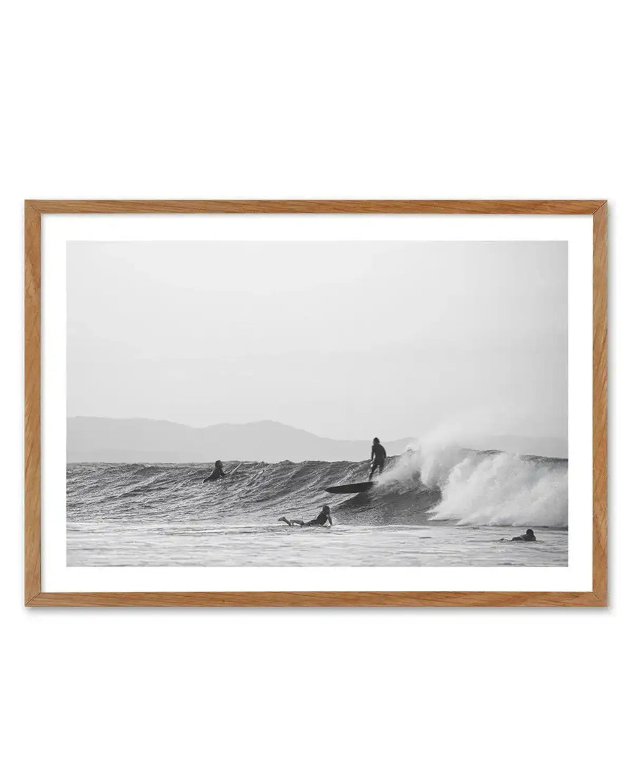 Framed black and white photograph of a coastal surf scene with a natural wood frame and white border. The composition features five surfers in the ocean, with one surfer riding a breaking wave, another paddling on a board, and three others waiting in the water. Mountains are visible in the distance under an overcast sky.