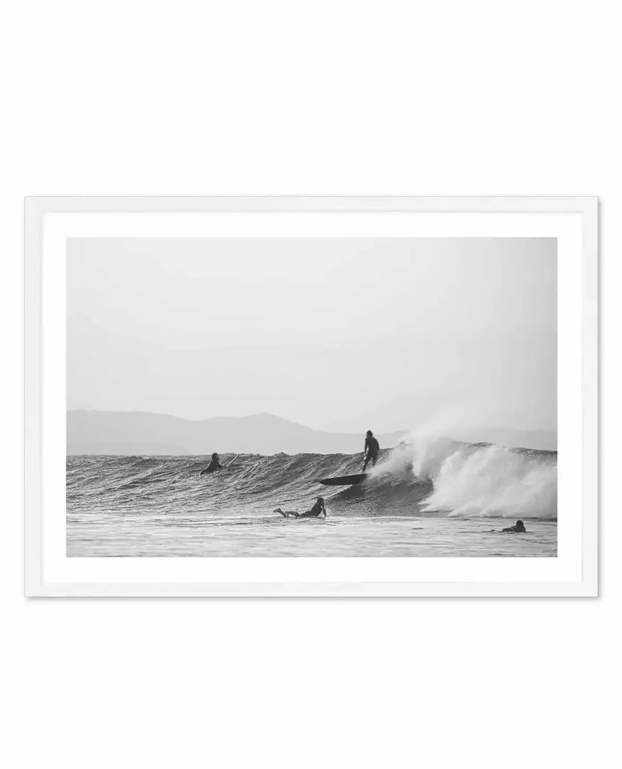 Framed black and white photograph of a coastal surf scene with a white frame and white border. The composition features five surfers in the ocean with a large wave breaking, and mountains in the background under an overcast sky.
