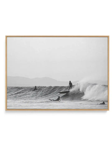 Black and white framed canvas print depicting multiple surfers in ocean waves with mountains visible in the hazy background. The photograph captures several figures in wetsuits at various points in the water—some paddling, one riding a large wave with white foam spray. The composition is horizontally oriented with a natural, documentary photography style. The canvas is mounted in a natural wood frame with a light tan finish.