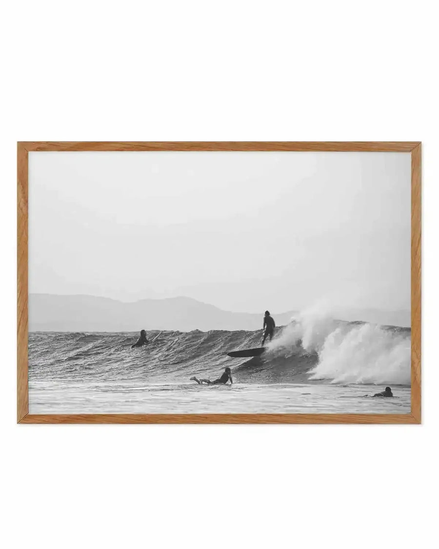 Framed black and white photograph of a surfing scene with a natural wood frame and white border. The composition features a surfer riding a breaking wave, with other surfers in the water and distant mountains under an overcast sky.