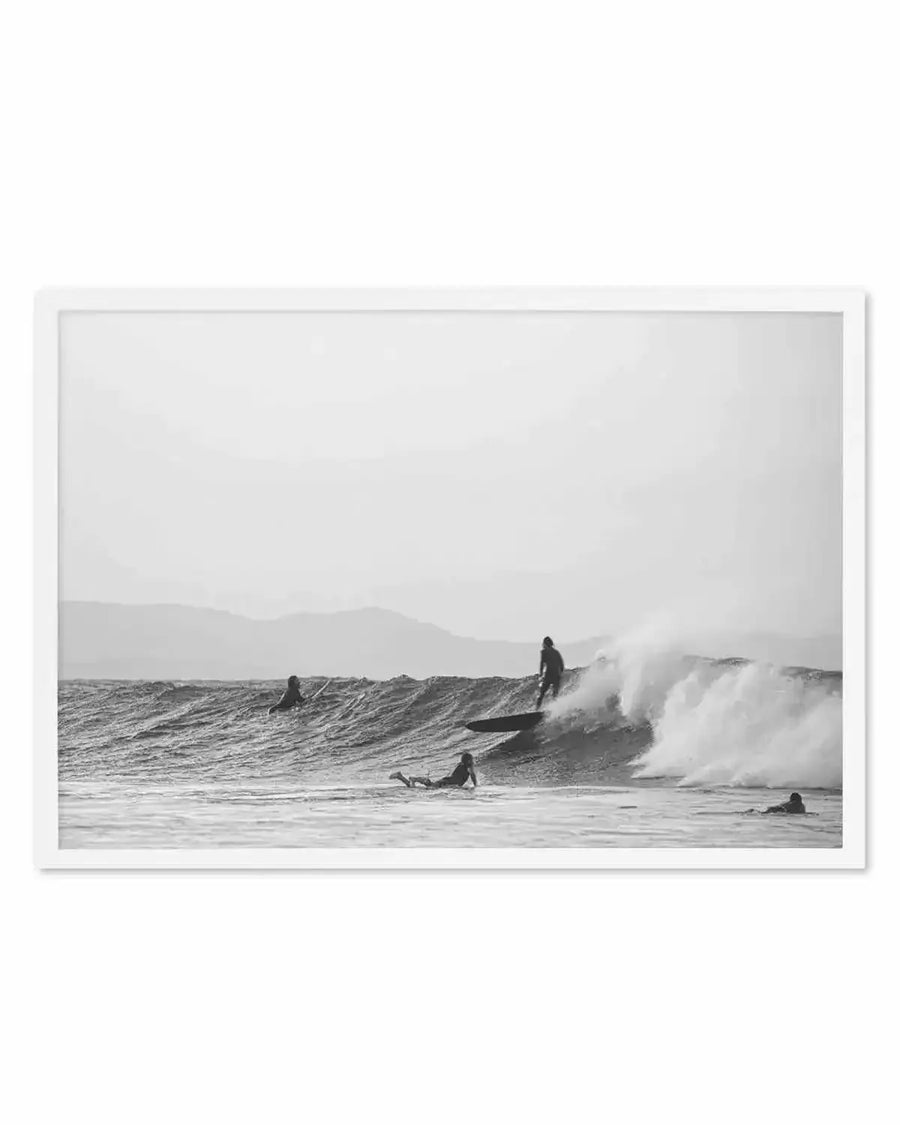 Framed black and white photograph of a dynamic ocean scene with surfers, displayed in a white frame with a white border. The composition captures a surfer riding a breaking wave, with other surfers waiting in the water and distant mountains under an overcast sky.