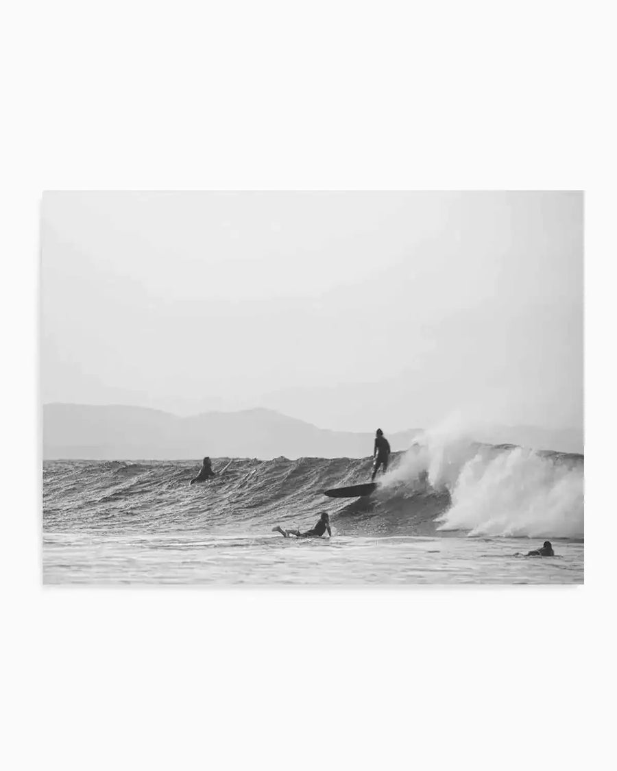 Framed black and white photograph of a coastal surf scene with a natural wood frame and white border. The composition features a surfer riding a wave with a large spray of water, while other surfers wait in the water. Mountains are visible in the background under an overcast sky.