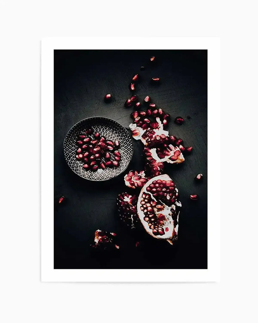 Framed photography art print featuring a top-down view of vibrant red pomegranate seeds and fruit pieces scattered across a dark, textured surface, with a black frame and white border. A small, patterned bowl filled with seeds is visible on the left.