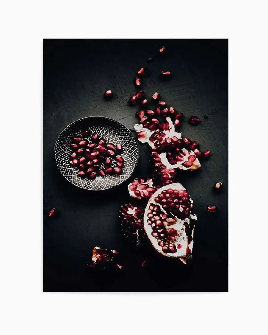 "Photography art print featuring a still life composition of pomegranates on a dark, textured surface. A small, patterned bowl holds a pile of ruby red pomegranate seeds, while a larger, partially opened pomegranate with visible seeds and white pith lies beside it. Scattered seeds and broken pieces of pomegranate are spread across the dark background, creating a rich, moody atmosphere with deep reds and blacks."