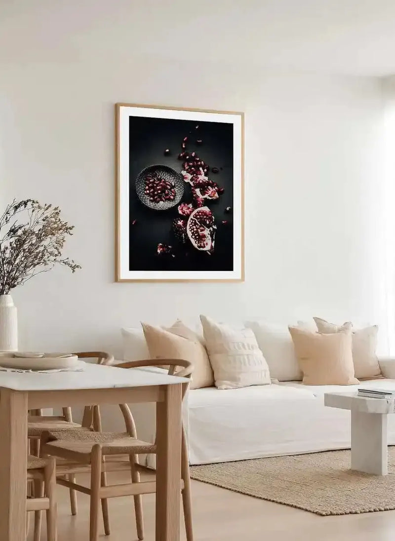 Large framed art print displayed above a white sofa in a minimalist living room. The print features a still life photograph of a halved pomegranate and loose pomegranate seeds in a small patterned bowl, scattered across a dark background, with a natural wood frame and white border. The room includes a white sofa with cream and beige cushions, a light wood dining table with chairs, and a white vase with dried branches.