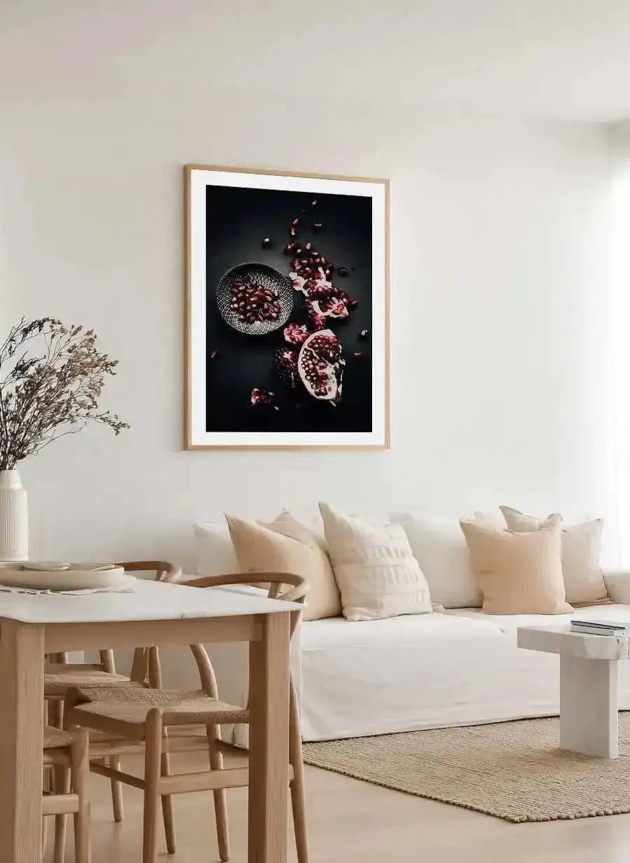 Large framed art print displayed above a white sofa in a minimalist living room. The print features a still life photograph of a halved pomegranate and loose pomegranate seeds in a small patterned bowl, scattered across a dark background, with a natural wood frame and white border. The room includes a white sofa with cream and beige cushions, a light wood dining table with chairs, and a white vase with dried branches.
