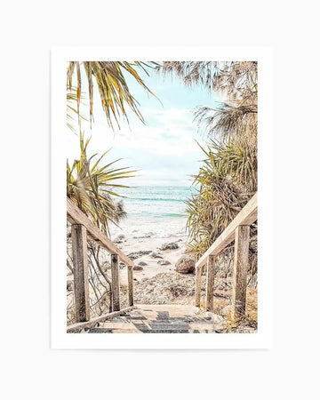 Framed photograph of a beach scene with natural wood frame and white border. The composition features a wooden staircase leading down to a sandy beach with gentle turquoise waves and a light blue sky. Lush green and golden-brown foliage frames the view, with scattered rocks on the sand.
