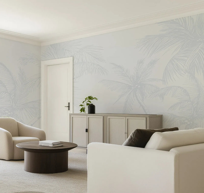 Lifestyle shot of a minimalist living room featuring The Palms Wallpaper in Duck Egg Blue. The wallpaper covers all visible walls with a repeating pattern of large-scale palm fronds and trees in a soft, muted duck egg blue against a light, almost white background. The room includes a cream-colored armchair, a round dark wood coffee table with books, and a white sofa with dark throw pillows. A light wood cabinet with closed doors and a dark vase with a green plant sits against the wallpapered wall.