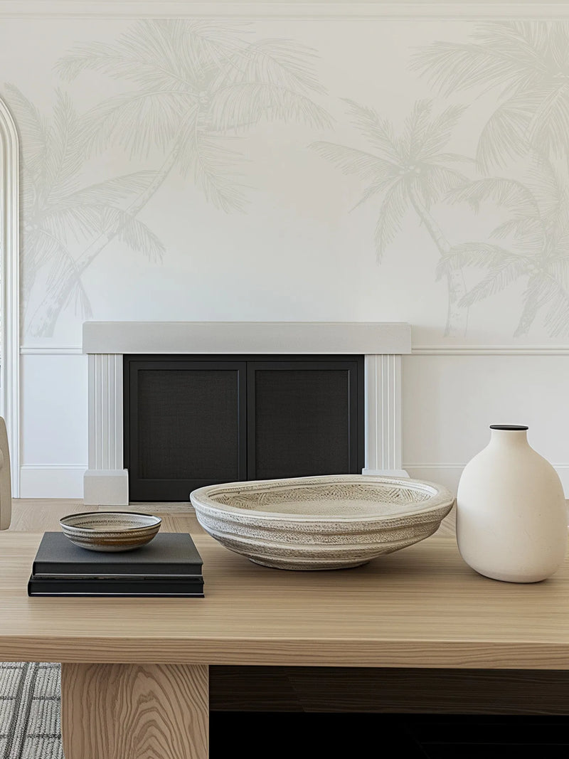 Lifestyle shot of a living room featuring The Palms Wallpaper in Grey. The wallpaper has a repeating pattern of hand-drawn palm trees in a soft, muted grey on a light cream background, creating a subtle tropical motif. The wallpaper is applied to a wall above a white fireplace with a black insert. In the foreground, a light wood coffee table displays a large textured cream bowl, a smaller striped bowl on top of two black books, and a cream ceramic vase.
