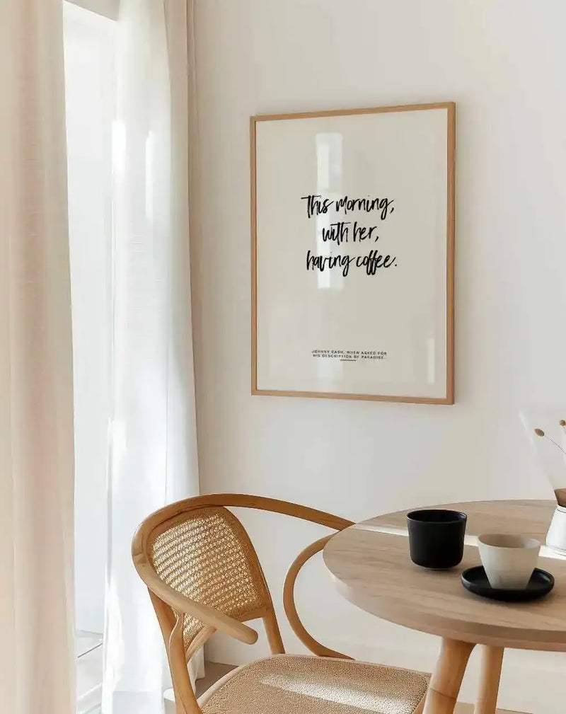 Lifestyle shot of a minimalist dining area featuring a framed art print above a round wooden table with two coffee cups. The print has a cream background with the phrase "This morning, with her, having coffee." in black script, framed in natural oak with a white border. A light wooden chair with a woven cane back is partially visible, and sheer white curtains hang by a window, letting in soft light.